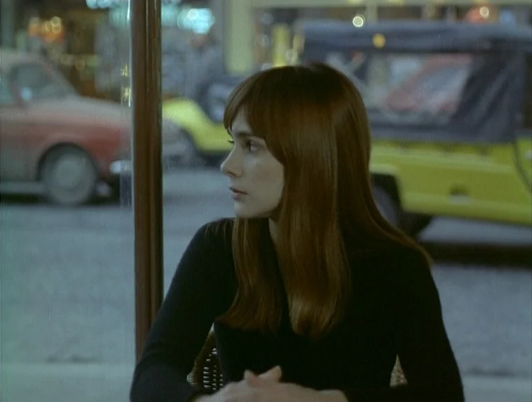 Love In The Afternoon (Chloe In The Afternoon) (L’amour l’après-midi) (1972) movie still: medium — A woman sits inside a dimly lit cafe behind a…