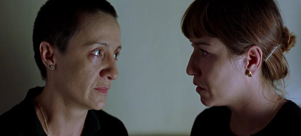 Volver (2006) movie still: medium — A dimly lit room with plain walls provides the backdrop for the two women. The women face each other in a…