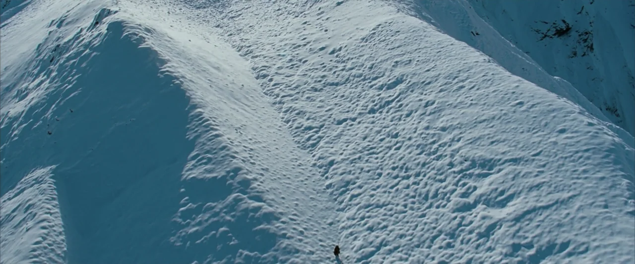 10,000 BC (2008) movie still: establishing — An aerial view looks down over a vast, snow-covered mountain slope. Deep furrows and undulations carve…