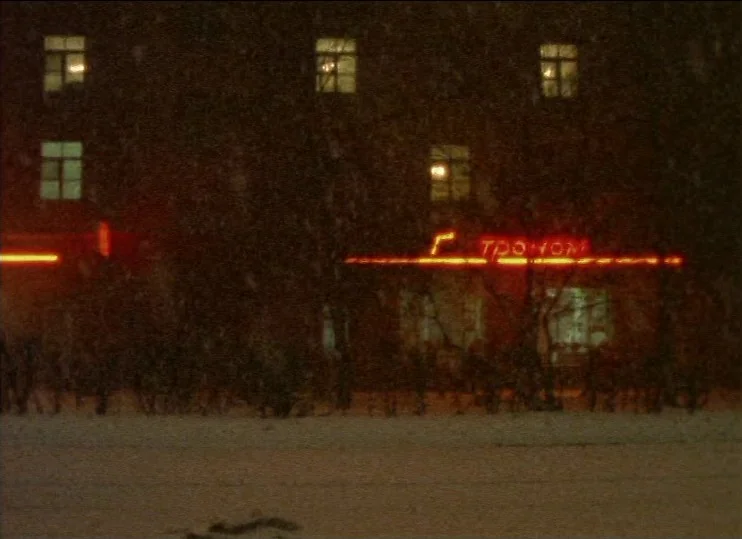 From The East (1993) movie still: establishing — A dark multi-story building at night with lit windows and a red neon sign. Snow falls between the…