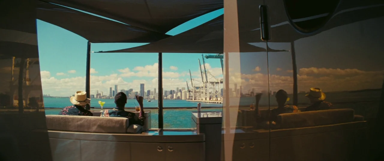 The Beach Bum (2019) movie still: wide — A modern yacht deck featuring sleek reflective surfaces and a canopy over the seating. Two individuals sit…