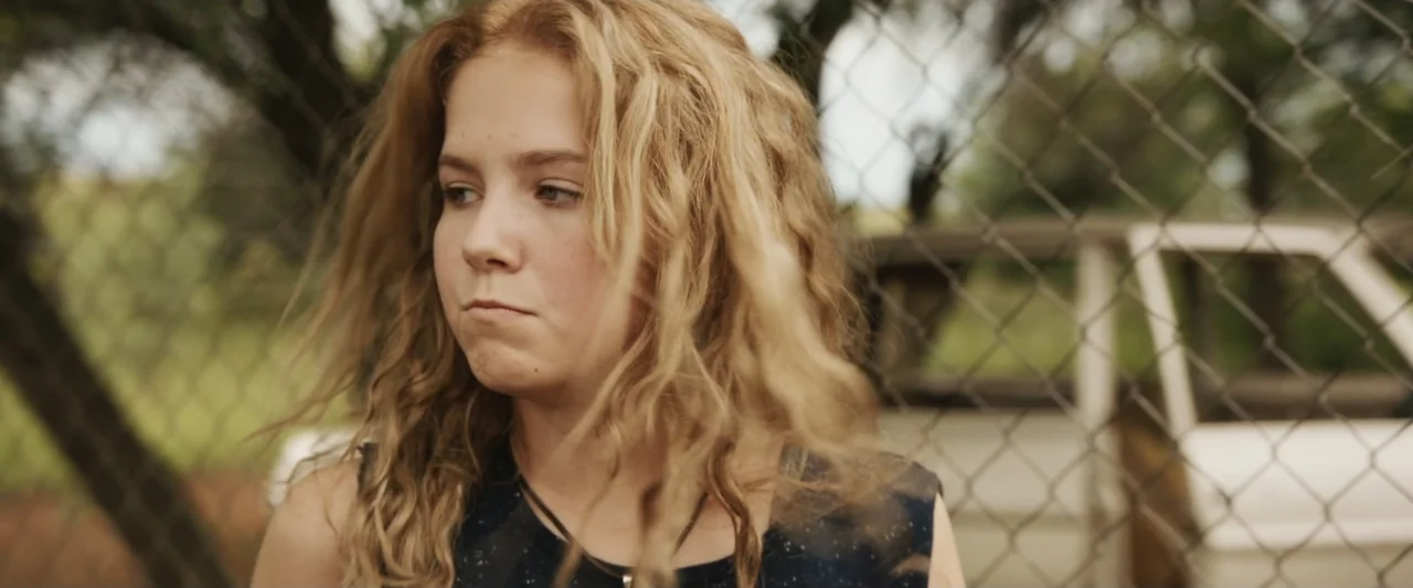 Mia and the White Lion (2018) movie still: medium — A teenager stands in front of a chain link fence outdoors during the day. She is oriented…
