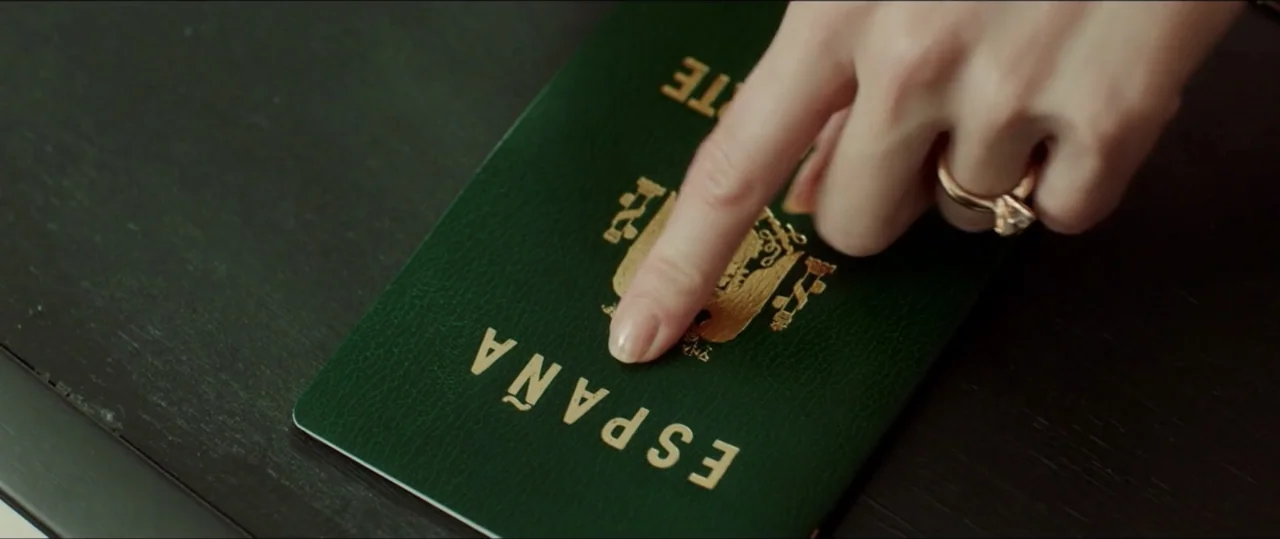 My Heart Goes Boom! (2020) movie still: closeup — A dark wooden surface holds a green Spanish passport with gold embossed lettering. A hand wearing…