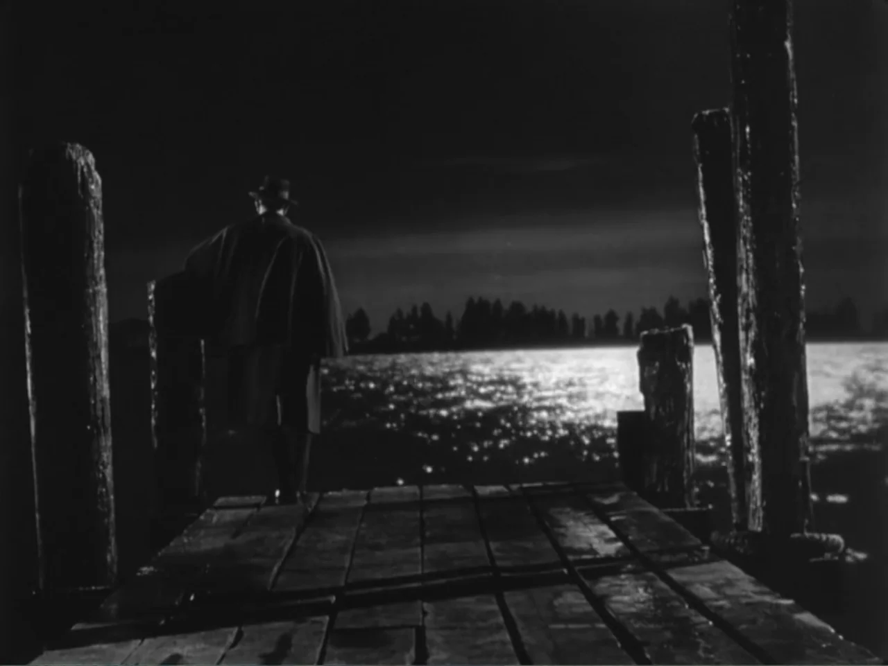 House by the River (1950) movie still: medium — A wooden pier extends into a dark body of water at night. A lone man in a trench coat and hat stands…