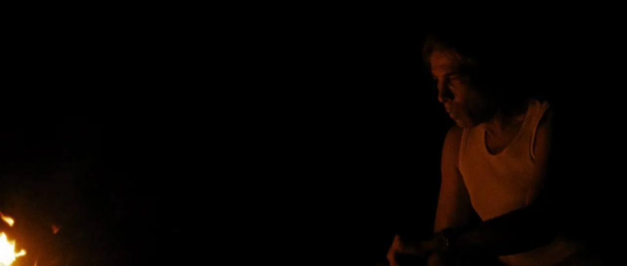 Let The Corpses Tan (2017) movie still: medium — A dark outdoor setting at night with a fire burning in the bottom left corner. A man sits by the…