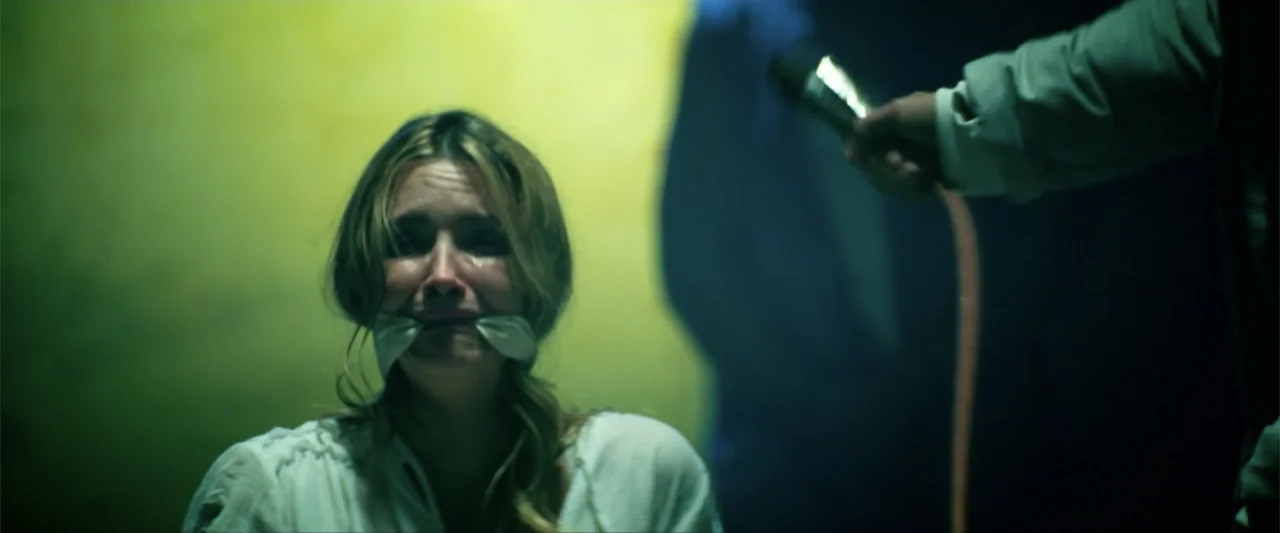 Bloodshot (2020) movie still: medium — A woman is centered in a dimly lit, yellow-walled room with a gag over her mouth. A blurred figure in the…