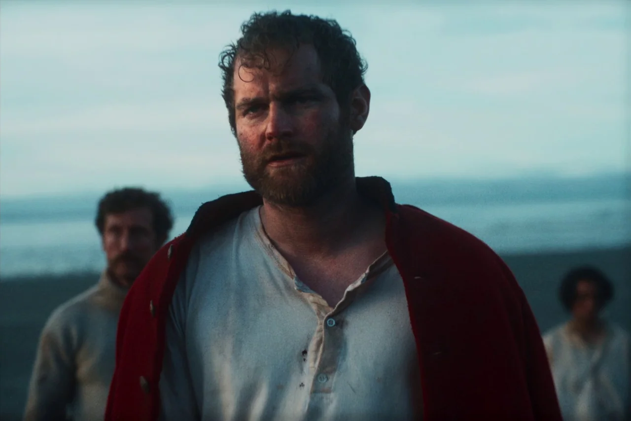 The Settlers (2023) movie still: medium — An overcast beach landscape at blue hour with sandy ground and distant water. A bearded man in a red coat…
