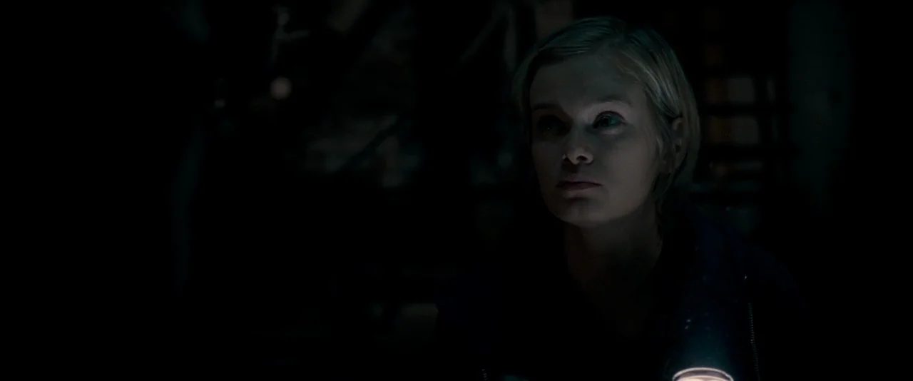 The Innkeepers (2011) movie still: closeup — A dark, underexposed environment with deep shadows concealing most of the room. A woman with blonde…