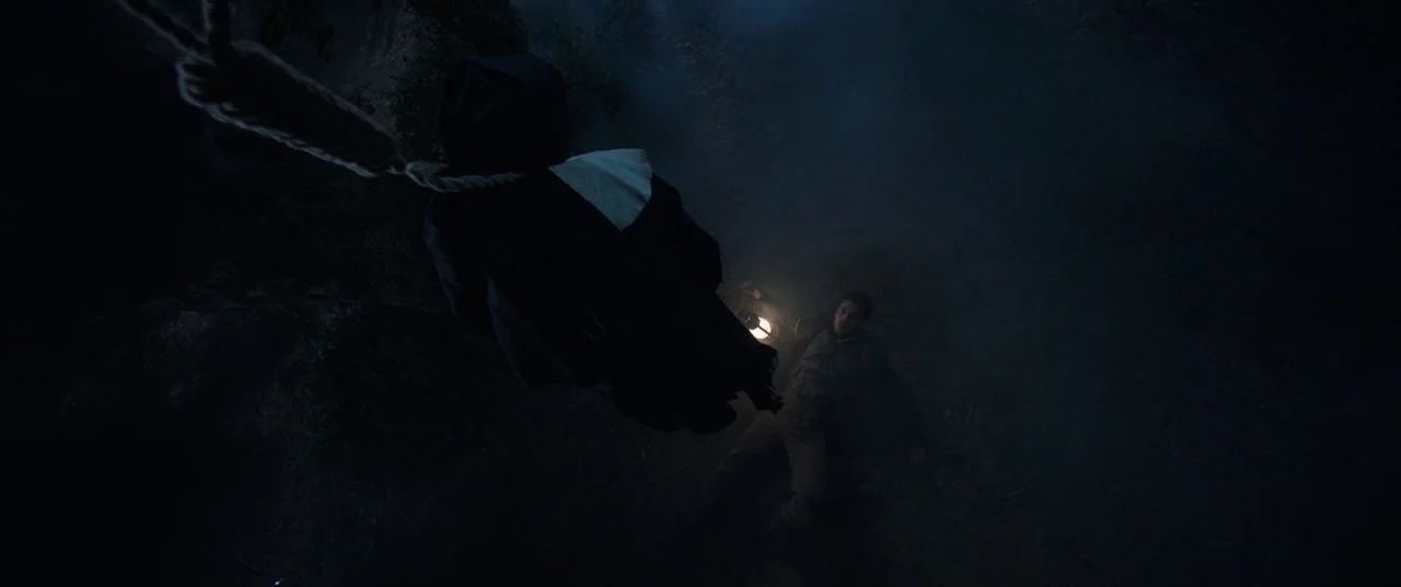 The Nun (2018) movie still: medium — A dark, misty, and uneven outdoor landscape at night with low visibility. A figure dressed as a nun hangs from…
