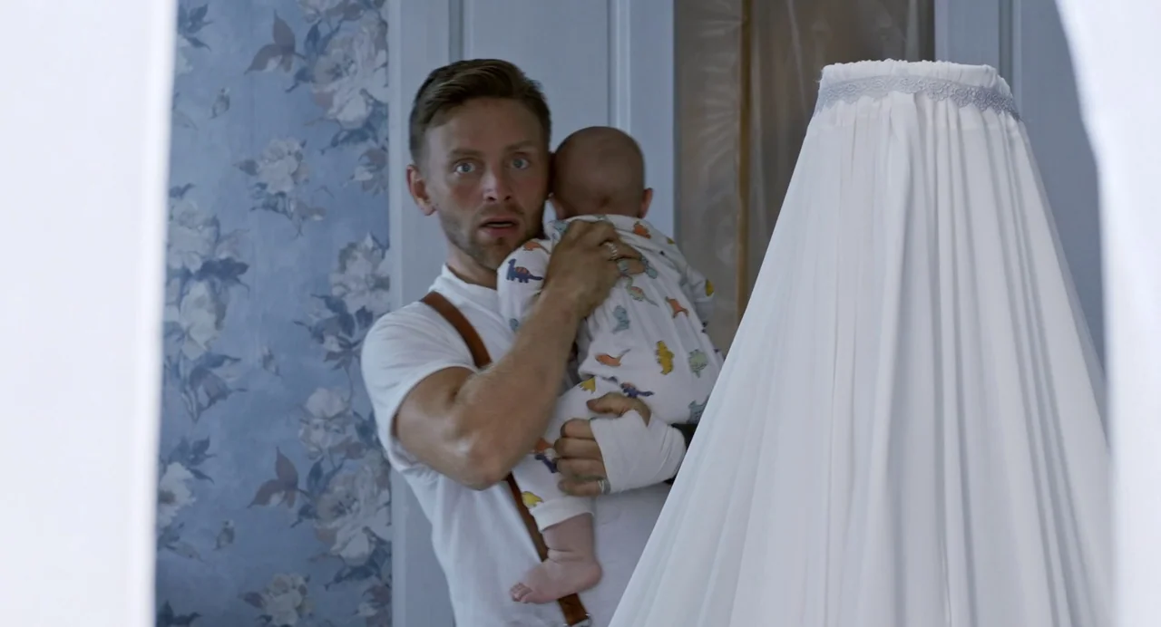 Hatching (2022) movie still: medium — A nursery features pale blue floral wallpaper and a draped white crib canopy. A man wearing suspenders holds…