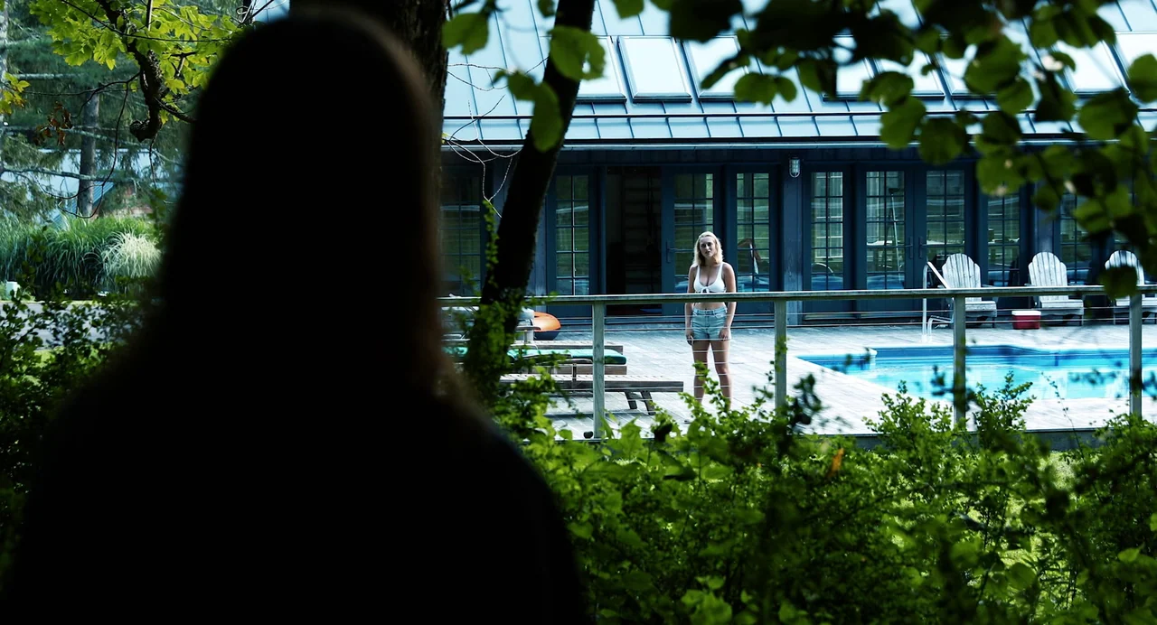 Hellbender (2021) movie still: wide — A modern dark-grey house with large glass doors faces a blue swimming pool deck. A young blonde woman in a…