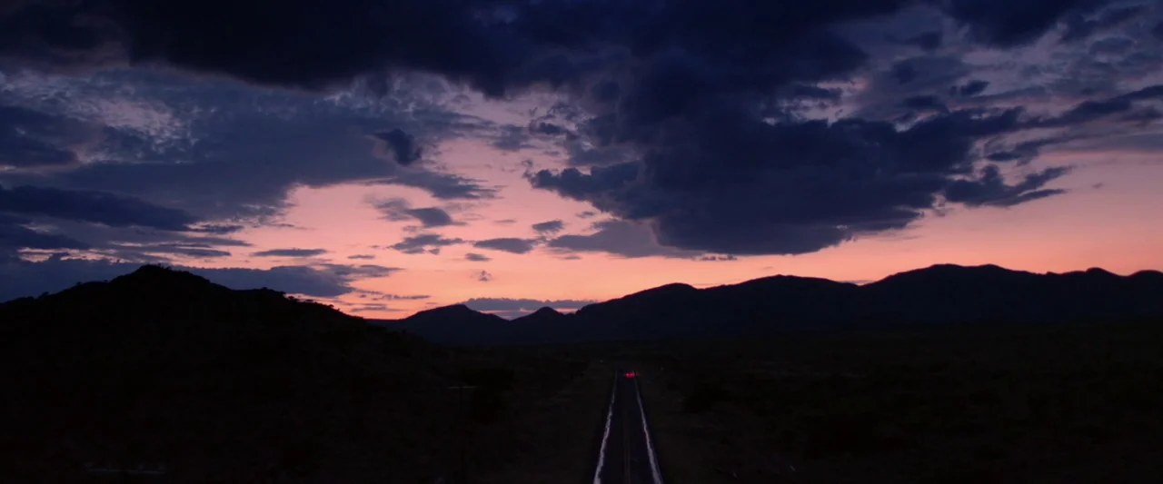 The Unknown Country (2022) movie still: establishing — A dark, expansive landscape features a distant mountain range under a colorful twilight sky.…