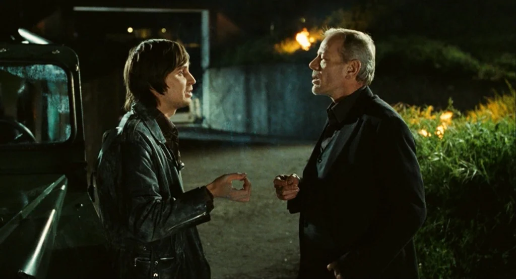This Night (2008) movie still: medium — A dark outdoor driveway at night features an unidentifiable vehicle to the left. Two men are standing near…