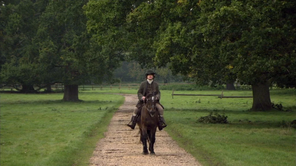 A Cock and Bull Story (2005) movie still: wide — A man rides a dark horse down a narrow gravel path between large trees. The rider wears period…
