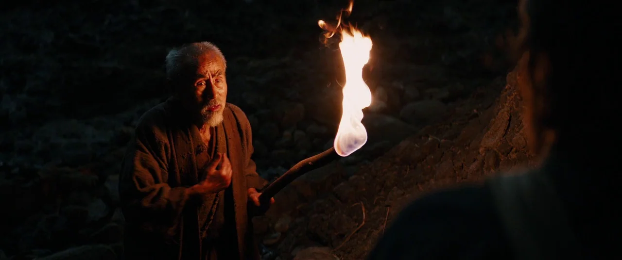 Silence (2016) movie still: medium — An elderly man stands in a dark, rocky outdoor area at night. He holds a brightly burning torch that…