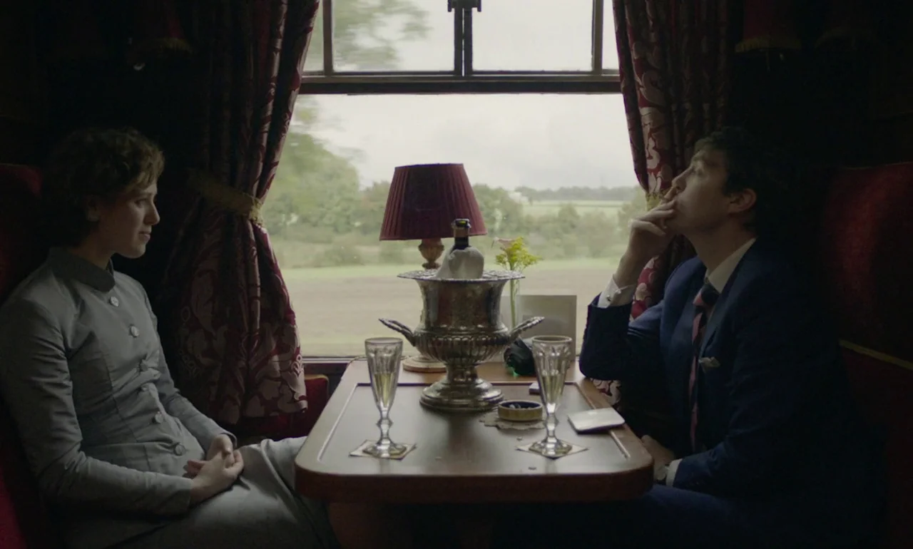 The Souvenir (2019) movie still: medium — A vintage railway cabin features red velvet curtains and wood paneling. A man and woman sit opposite each…