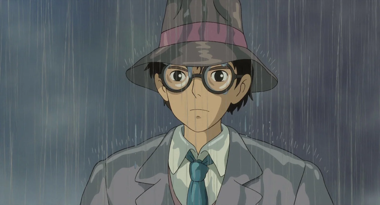 The Wind Rises (2013) movie still: medium — A man stands outdoors under heavy rainfall on an overcast day. He wears a grey fedora and an overcoat…