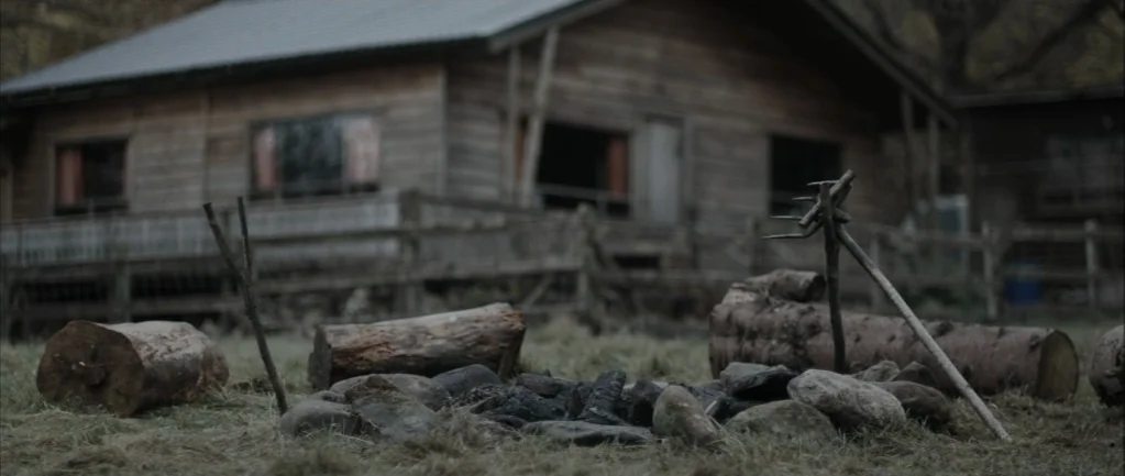 One Hundred Mornings (2009) movie still: wide — A stone-lined fire pit sits in a grassy yard in front of a wooden cabin. Several logs are arranged…