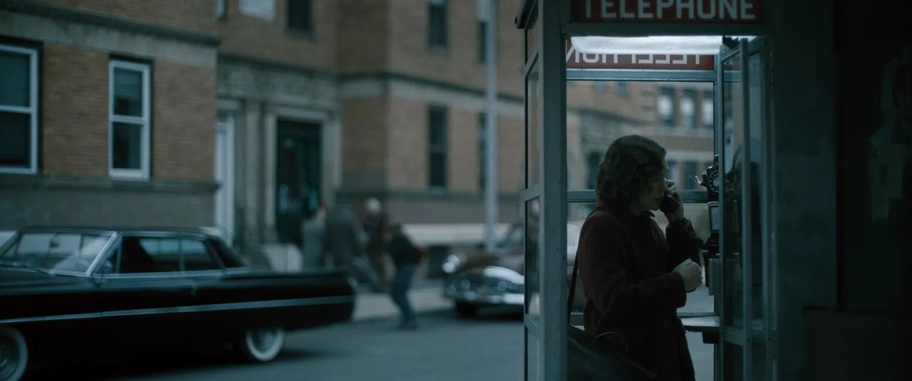 Boston Strangler (2023) movie still: medium — A street scene in a mid-century urban area with vintage cars parked nearby. A woman is inside a glass…
