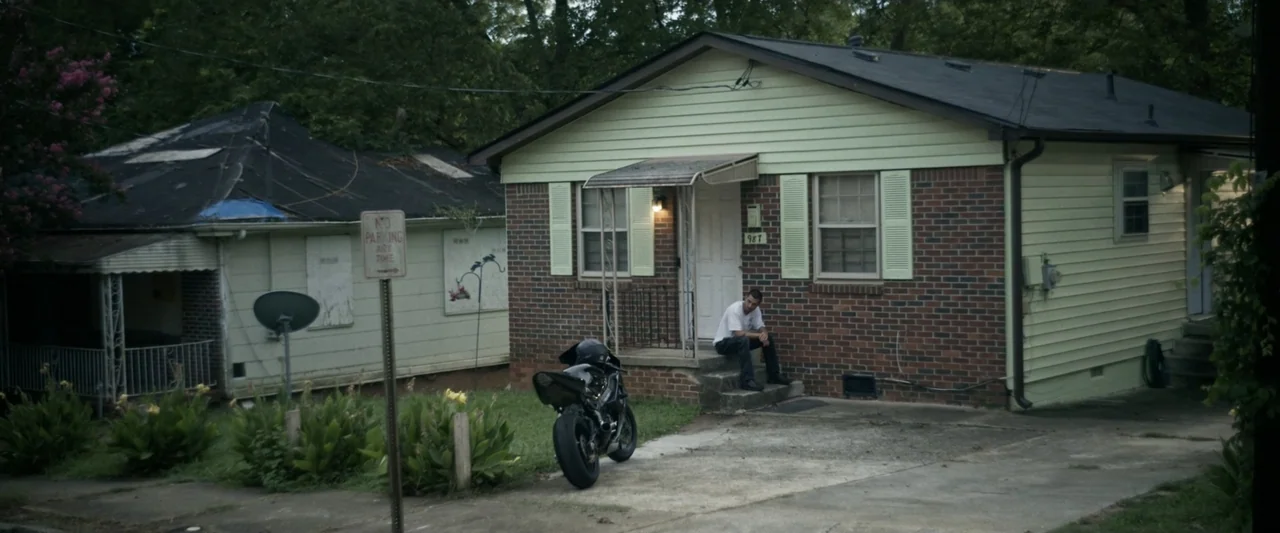 One Fast Move (2024) movie still: wide — A modest single-story home features light green siding and a brick-faced facade. A man sits alone on the…