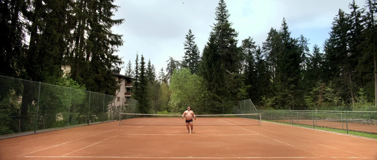 Youth (2015) movie still: wide — An outdoor clay tennis court is surrounded by a dense forest of evergreen trees. A shirtless man stands alone in…