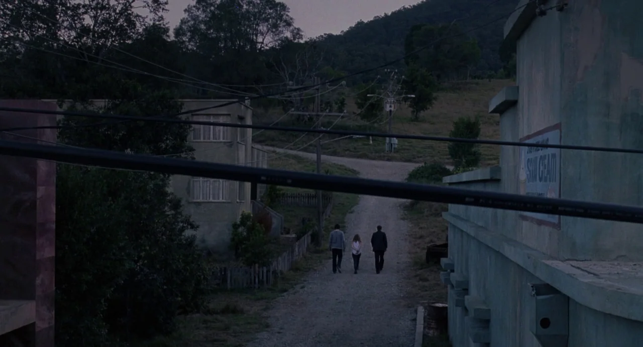 House of Wax (2005) movie still: wide — A quiet dirt road stretches toward a small house and a grassy hill at dusk. Three people walk away from the…