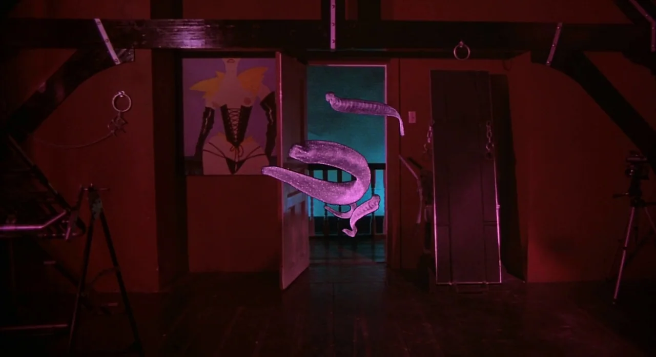 From Beyond (1986) movie still: wide — A dimly lit wooden attic is filled with red light and contains hanging shackles. Glowing, translucent,…