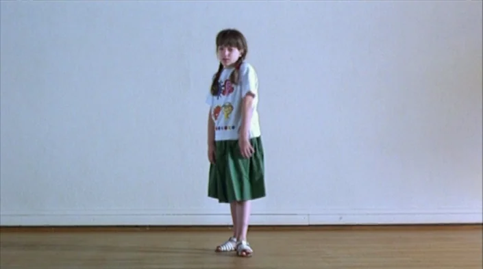 Code Unknown (2000) movie still: fullbody — A spacious room features plain white walls and a light-toned wooden floor. A young girl stands alone in…