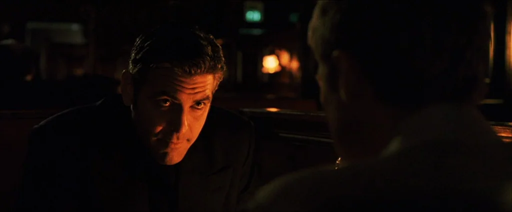 Ocean’s Eleven (2001) movie still: medium — A dimly lit interior with dark wooden furniture and soft ambient amber glow. A man in a dark suit sits…