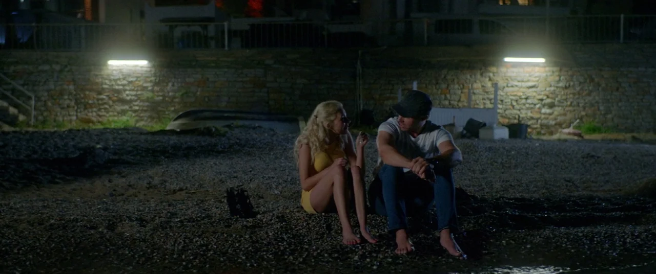 Holiday movie still: wide — A dark rocky beach sits at the base of a low stone retaining wall. A man and a woman sit together on the pebbles near…