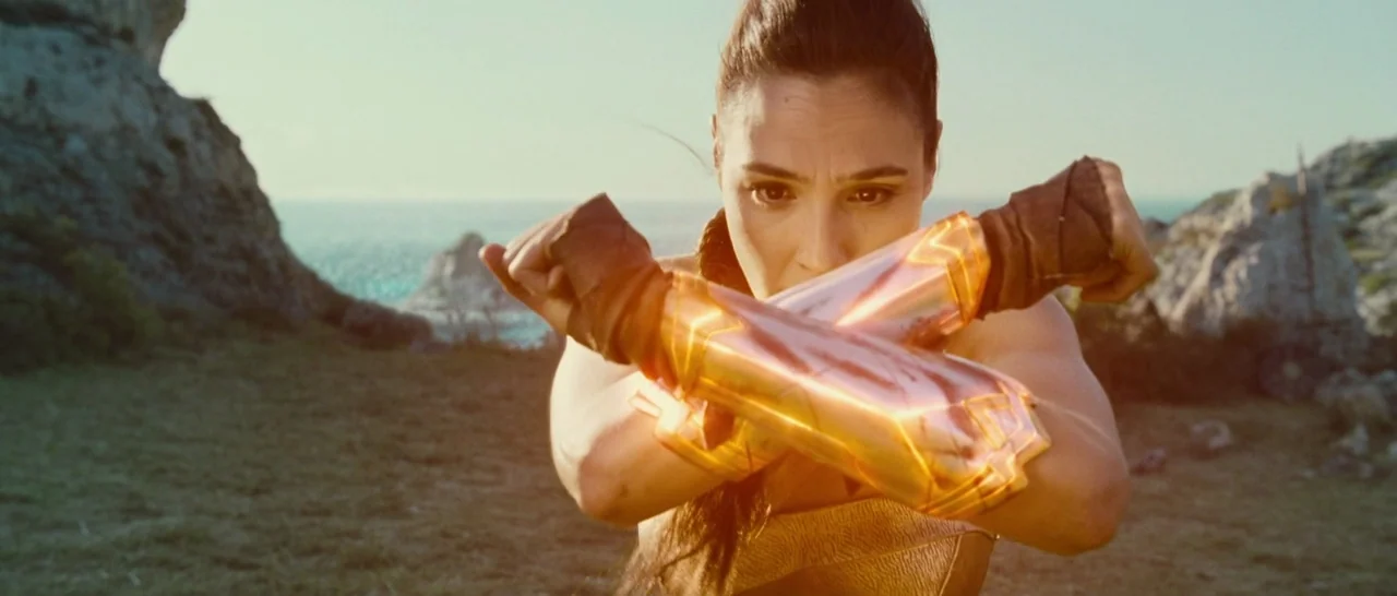 Wonder Woman (2017) movie still: medium — A grassy coastal cliff overlooks a sparkling blue ocean under a bright day sky. A woman wears metallic…