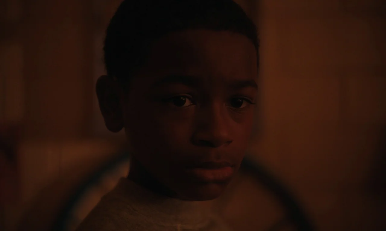 We Grown Now (2023) movie still: closeup — A dimly lit room provides a dark background for the close-up portrait. The subject looks forward with an…