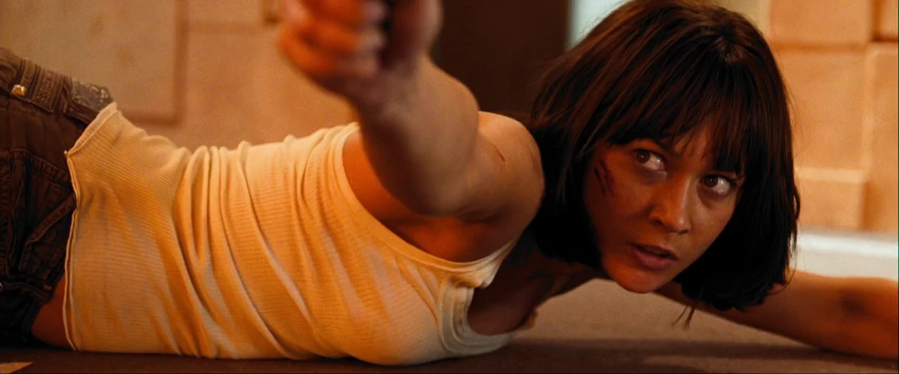 Quantum of Solace (2008) movie still: medium — A dimly lit indoor space with tiled walls. A woman lies prone on the ground while aiming a firearm…