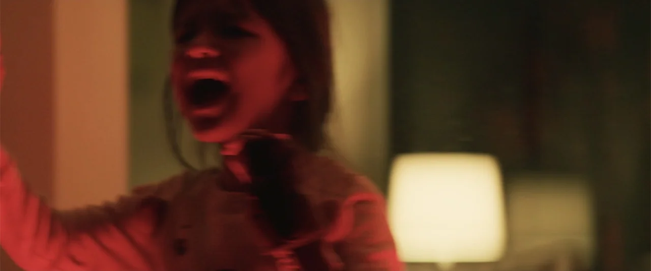 Human Animals (2020) movie still: closeup — A dimly lit room contains a blurred lamp in the background. A child stands in the foreground bathed in…
