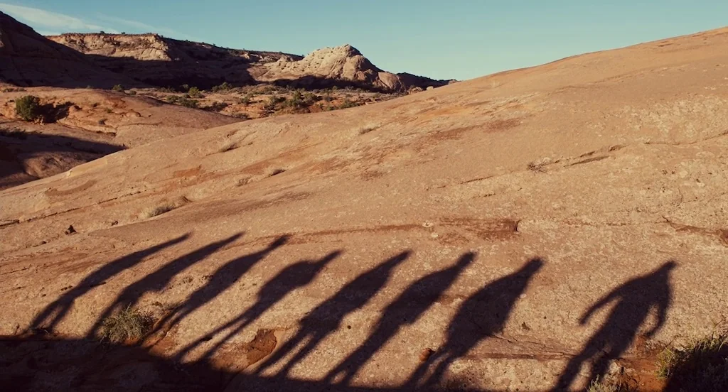 127 Hours (2010) movie still: wide — A vast, uneven expanse of orange-hued sandstone rock covers the frame under clear sky. Seven distinct,…
