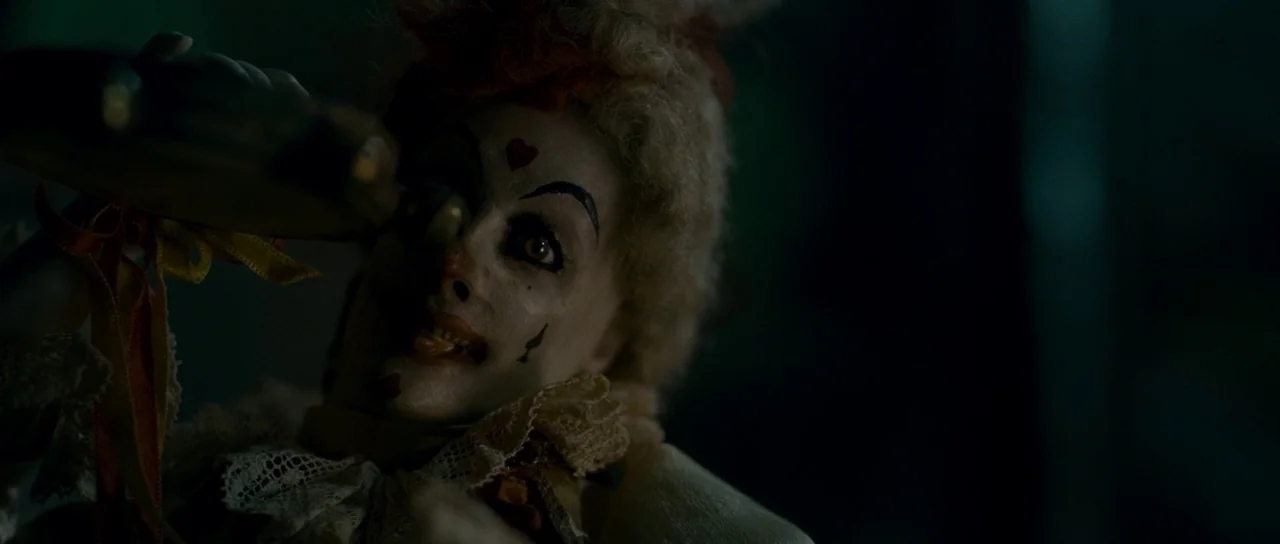 The Woman in Black (2012) movie still: closeup — A close-up view of a vintage doll with clown-like features in a dark environment. The doll looks…