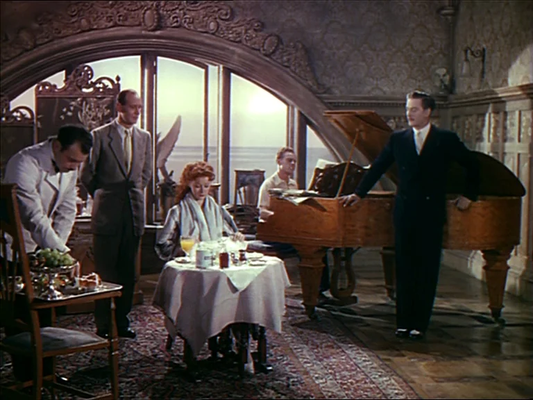 The Red Shoes (1948) movie still: wide — An ornate hotel room features an arched window overlooking the ocean and wood-paneled walls. Five people…