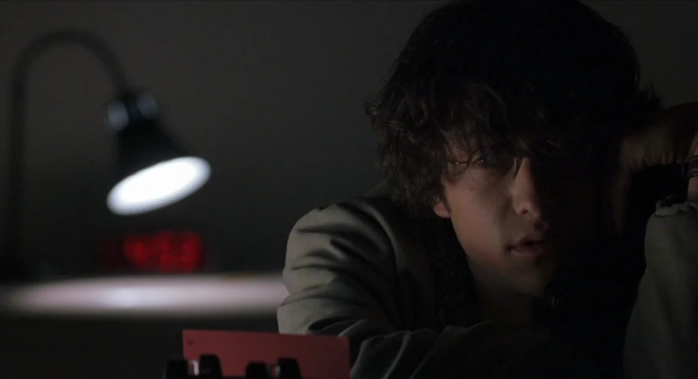 Brick (2005) movie still: closeup — A dimly lit room features a dark background with a glowing digital clock. A young man with glasses sits at a…