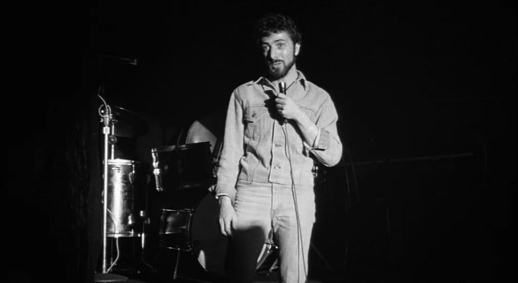 Lenny (1974) movie still: medium — A man stands on a dark stage with a drum kit behind him. He holds a microphone near his mouth and faces the camera.