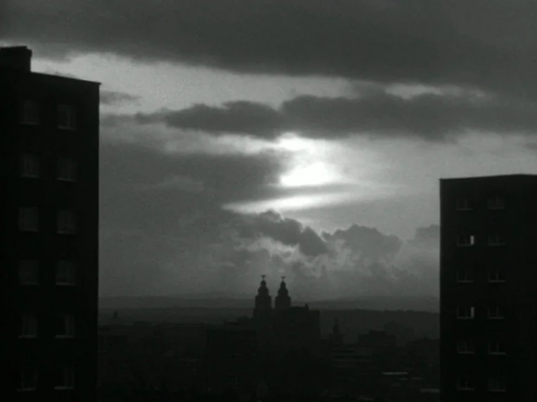 Death and Transfiguration (1983) movie still: establishing — A city skyline is visible in the distance beneath a bright, cloudy sky at dusk. Two…