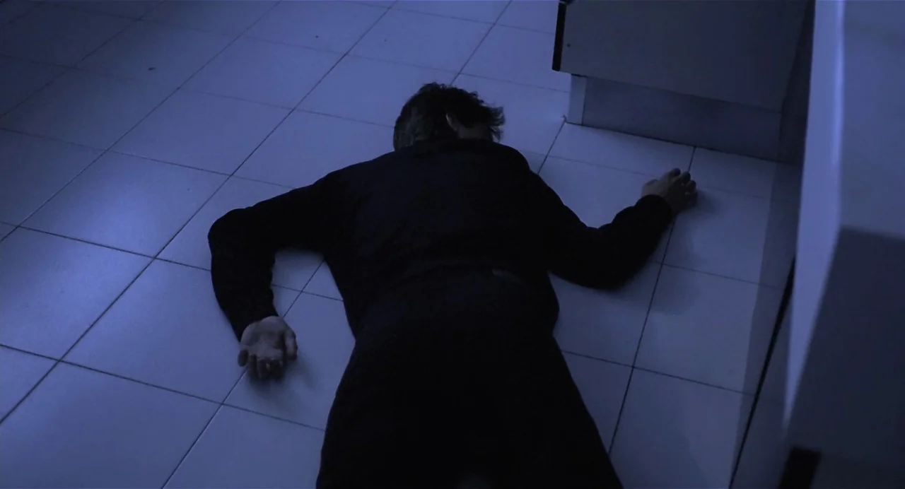 Thesis (1996) movie still: medium — A person lies face down on a square tiled floor in low light. The subject is wearing dark clothing and remains…