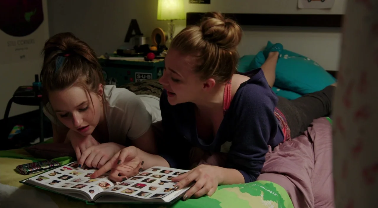 Laggies (2014) movie still: medium — A dimly lit room with a dark wooden bed frame and a green lamp. Two teenage girls lie on the mattress while…