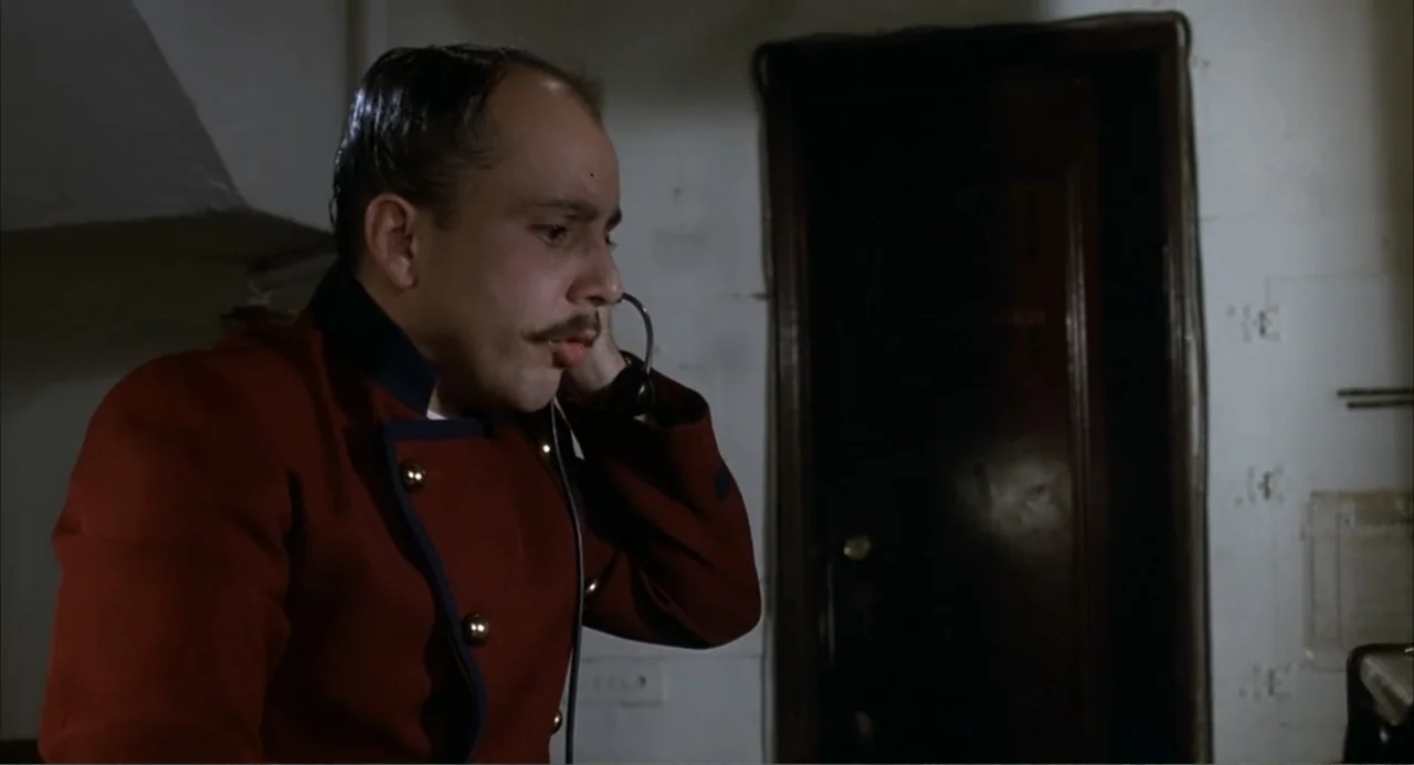 I, Madman (1989) movie still: medium — A dimly lit hotel hallway with a single dark door visible in the background. A man in a red uniform stands in…