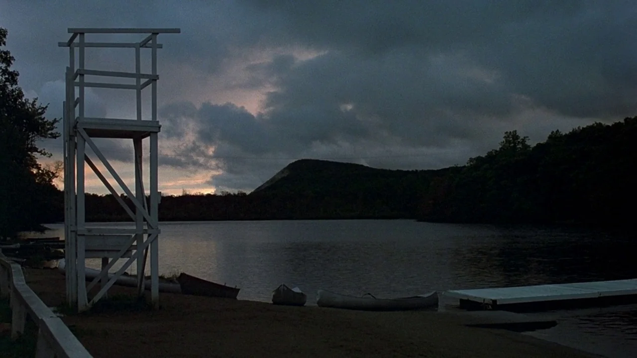 Friday the 13th (1980) movie still: establishing — A dark lakeside scene at dusk features a white lifeguard chair and canoes. A large silhouetted…