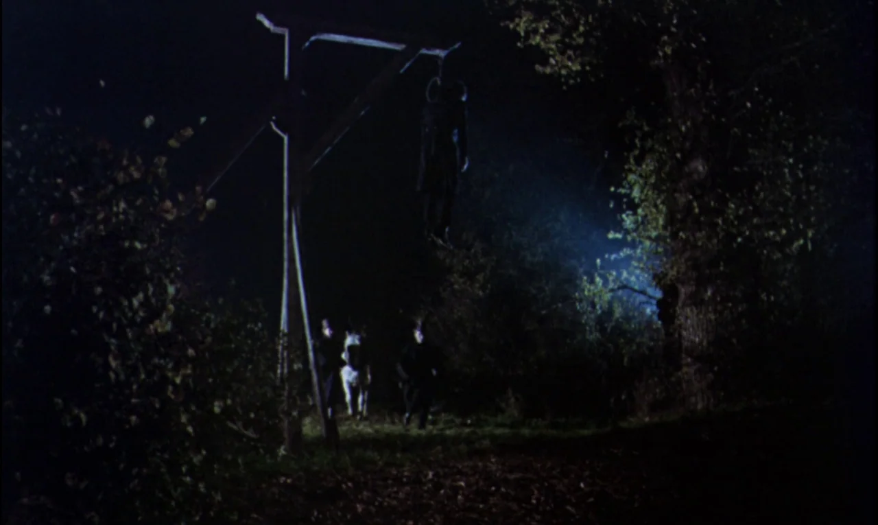 The Curse of Frankenstein (1957) movie still: wide — A dark night landscape featuring a wooden gallows structure erected in a wooded area. A body…