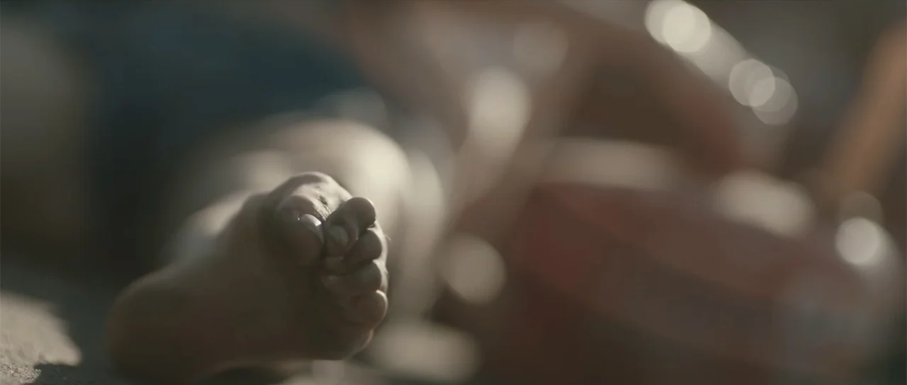 Night of the Missing (2023) movie still: closeup — A close view of a human foot resting on a sandy beach surface. The background is blurred with…