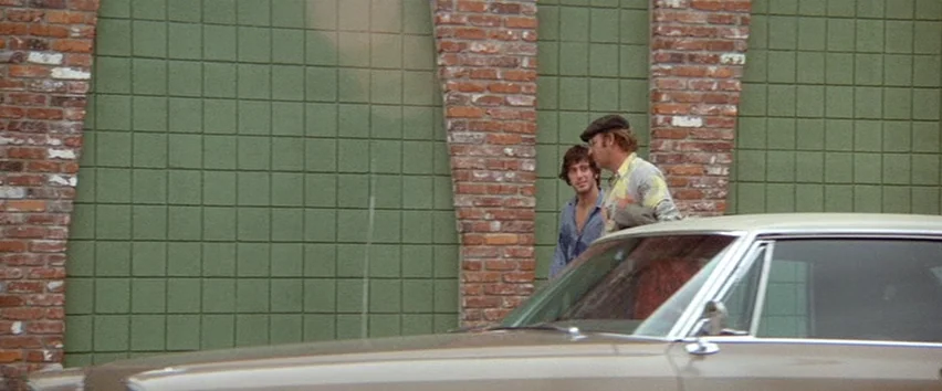 Scarecrow (1973) movie still: medium — Two men stand together before a brick wall with green tile accents. They pause near a parked sedan while…