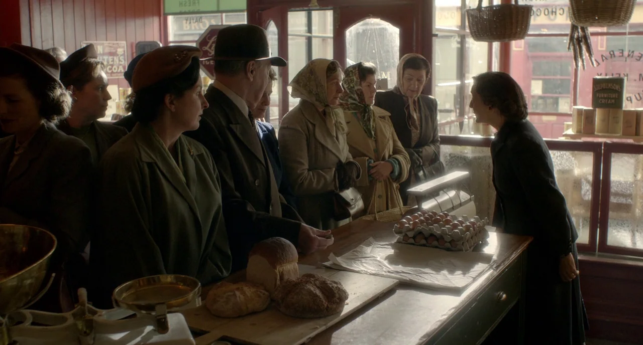 Brooklyn (2015) movie still: medium — A 1940s general store interior features a wooden counter laden with bread and eggs. A line of customers in…
