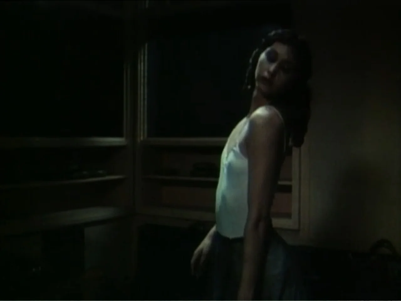Curtains (1995) movie still: medium — A dark bedroom interior features built-in shelves against a dimly lit back wall. A young woman in a white tank…
