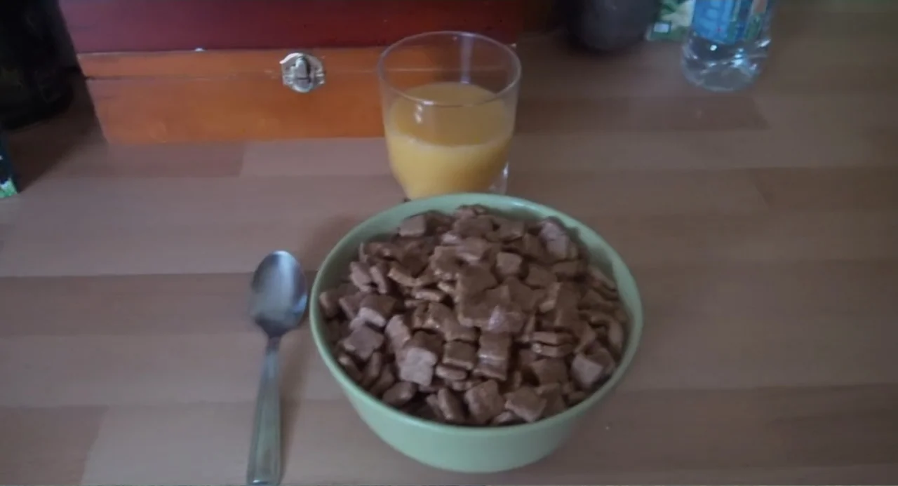 A Dramatic Film (2019) movie still: medium — A light wood table holds a bowl of cereal, a glass of juice, and a spoon. The items sit arranged for a…